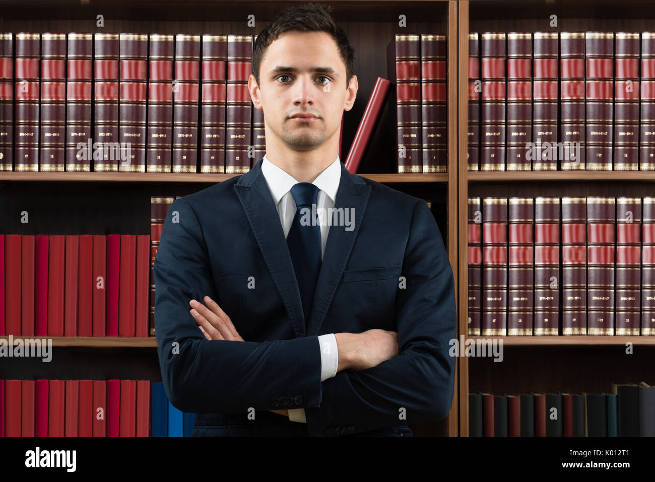 Ritratto di fiducia avvocato maschio in piedi con le braccia incrociate contro scaffale in aula Foto Stock