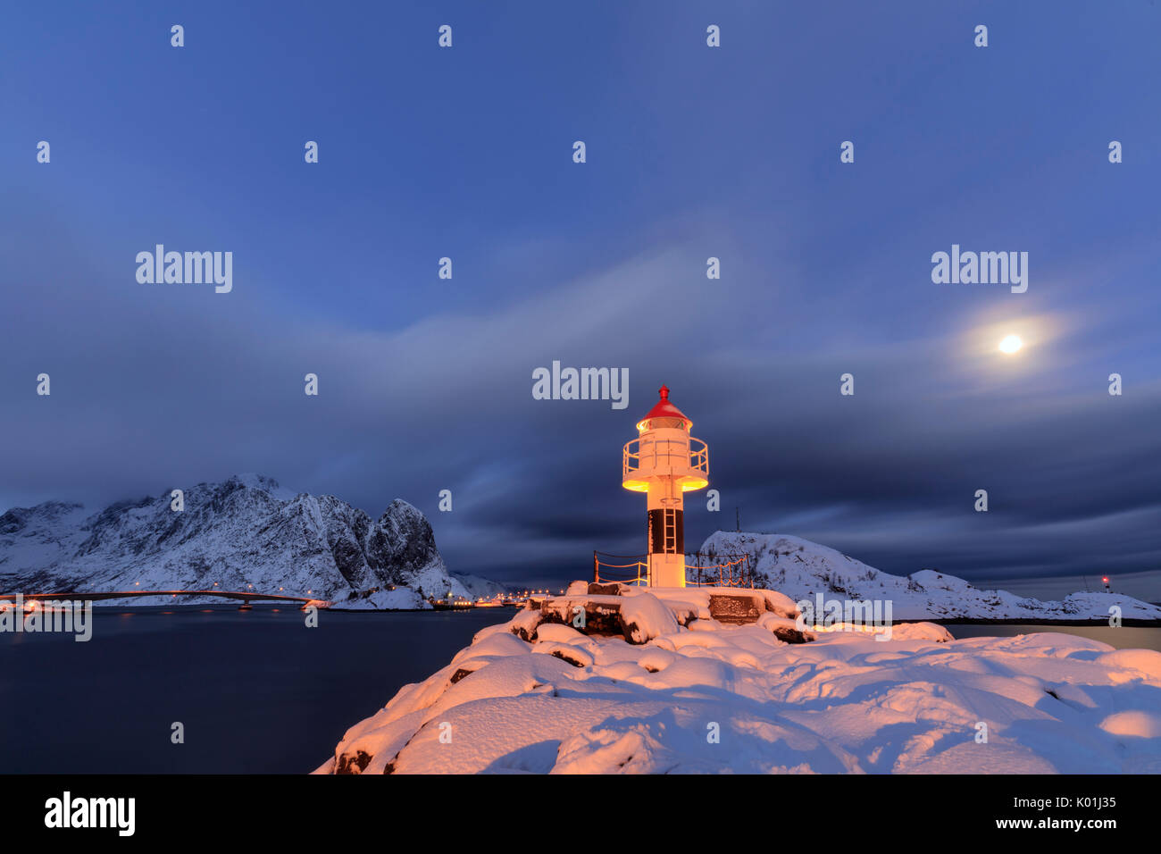 Faro e luna piena nella notte artica con il villaggio di Reine in background Nordland Isole Lofoten in Norvegia Europa Foto Stock