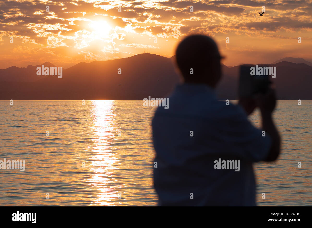 Il lago di Garda, Italia, uomo di scattare una foto del paesaggio lacustre al tramonto con un telefono cellulare fotocamera Foto Stock