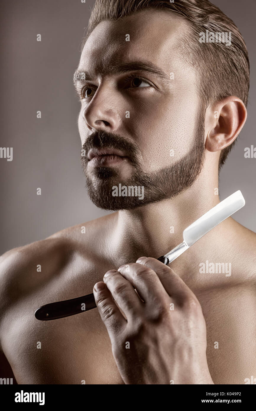 Primo piano del giovane uomo bello ragazzo con la barba che mostra una lama di rasoio, studio shot. Concetto di rasatura Foto Stock