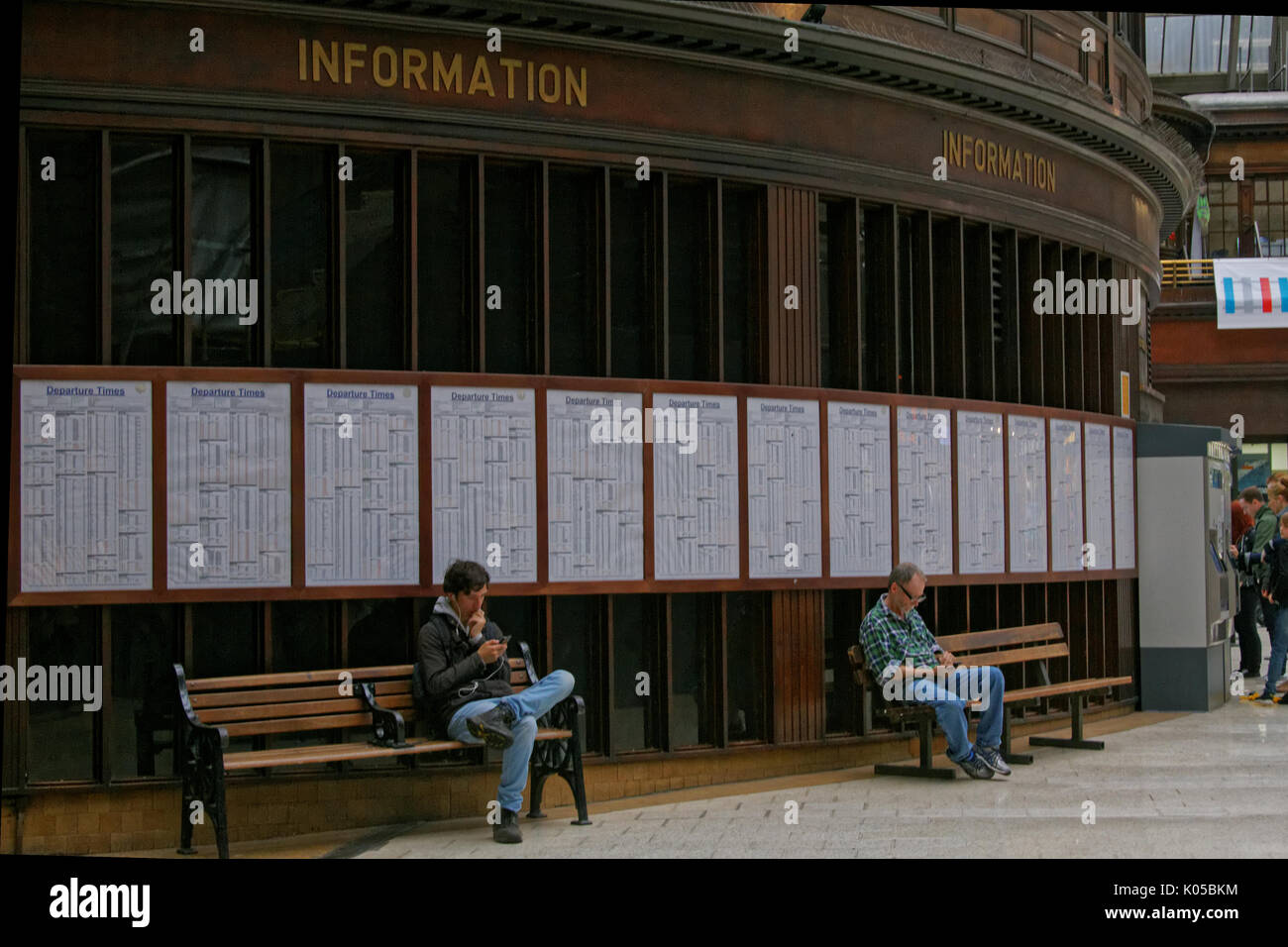 Due uomini ragazzi seduti sui banchi del treno tabelle tempo sfondo utilizzando smart phone sotto in pannelli di legno segno informazioni la stazione centrale di Glasgow concourse Foto Stock