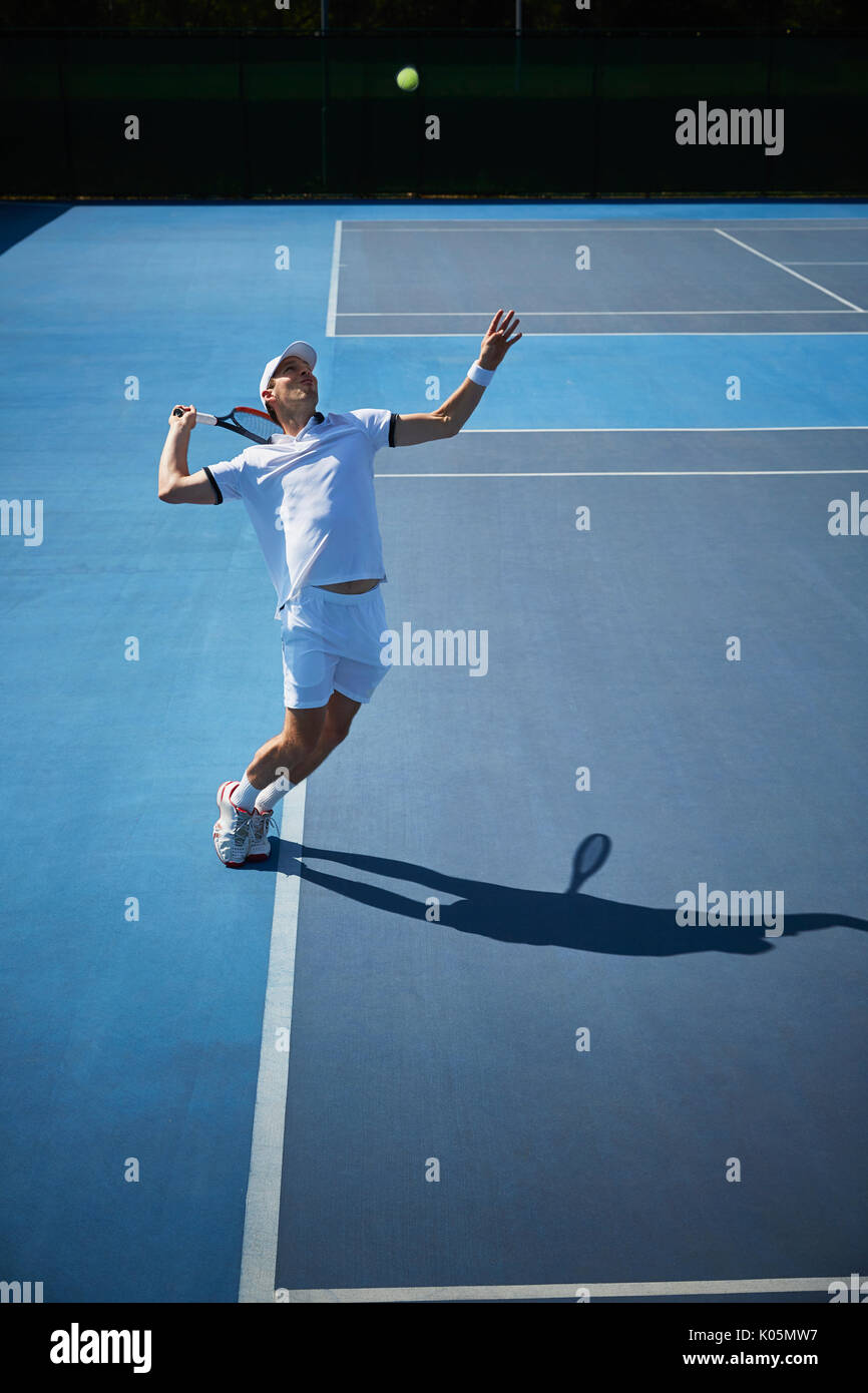 Maschio giovane giocatore di tennis giocando a tennis, che serve la sfera sul blu soleggiato Campo da tennis Foto Stock