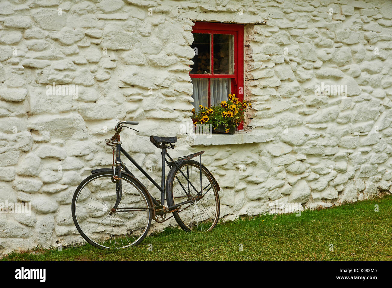 Una vecchia bicicletta appoggiata contro un lime lavato costruzione in pietra con un dipinto di rosso finestra e fiori di colore giallo Foto Stock