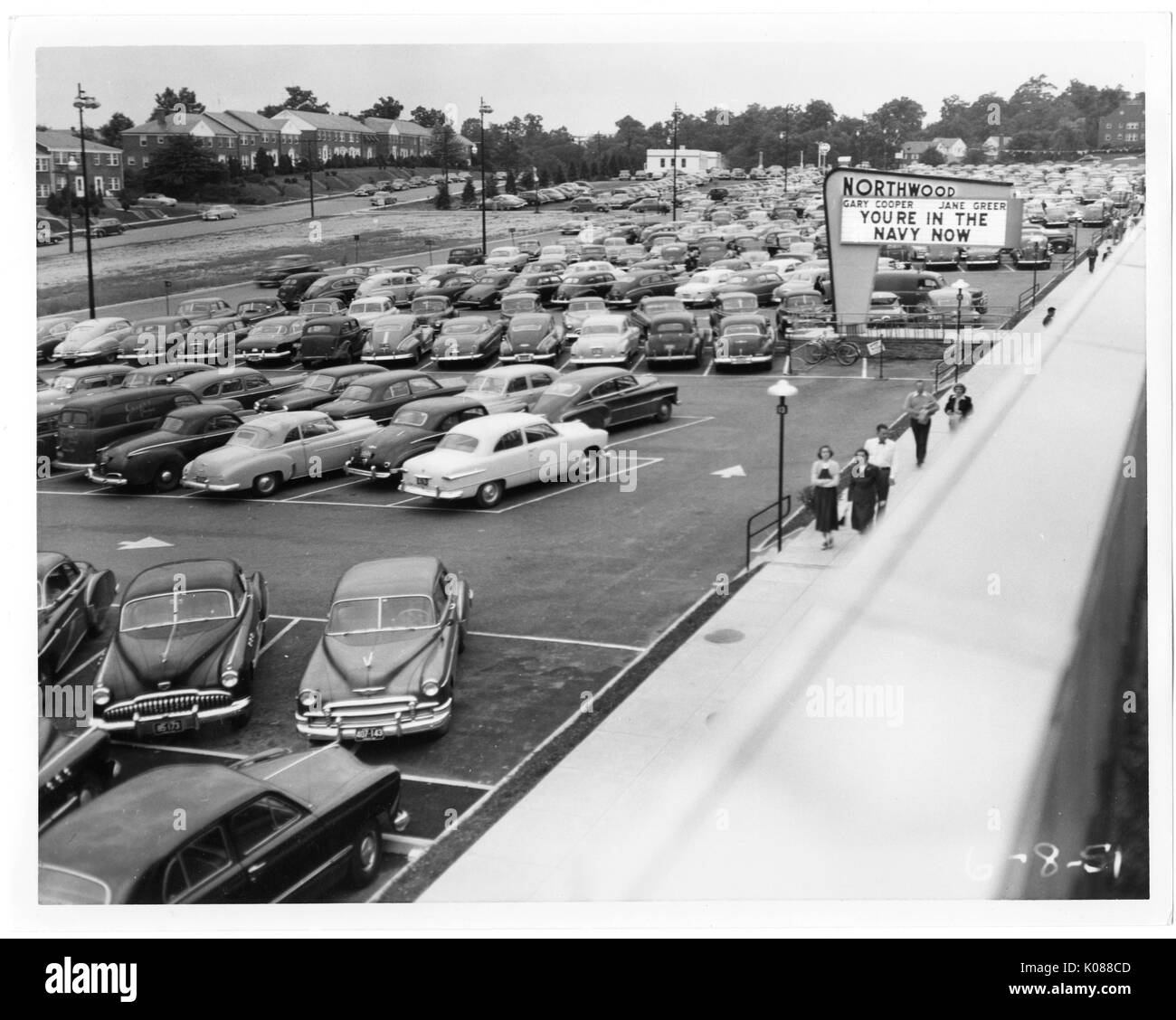 Vista di un pranzo parcheggio dal tetto del Northwood Shopping Centre, attraversata la strada dal centro e il parcheggio sono fila di mattoni case, molte persone stanno camminando sul marciapiede del centro del centro è di segno pubblicità 'YOURE IN MARINA ORA' con 'Gary Cooper " e " Jane Greer', Baltimore, Maryland, 1951. Foto Stock