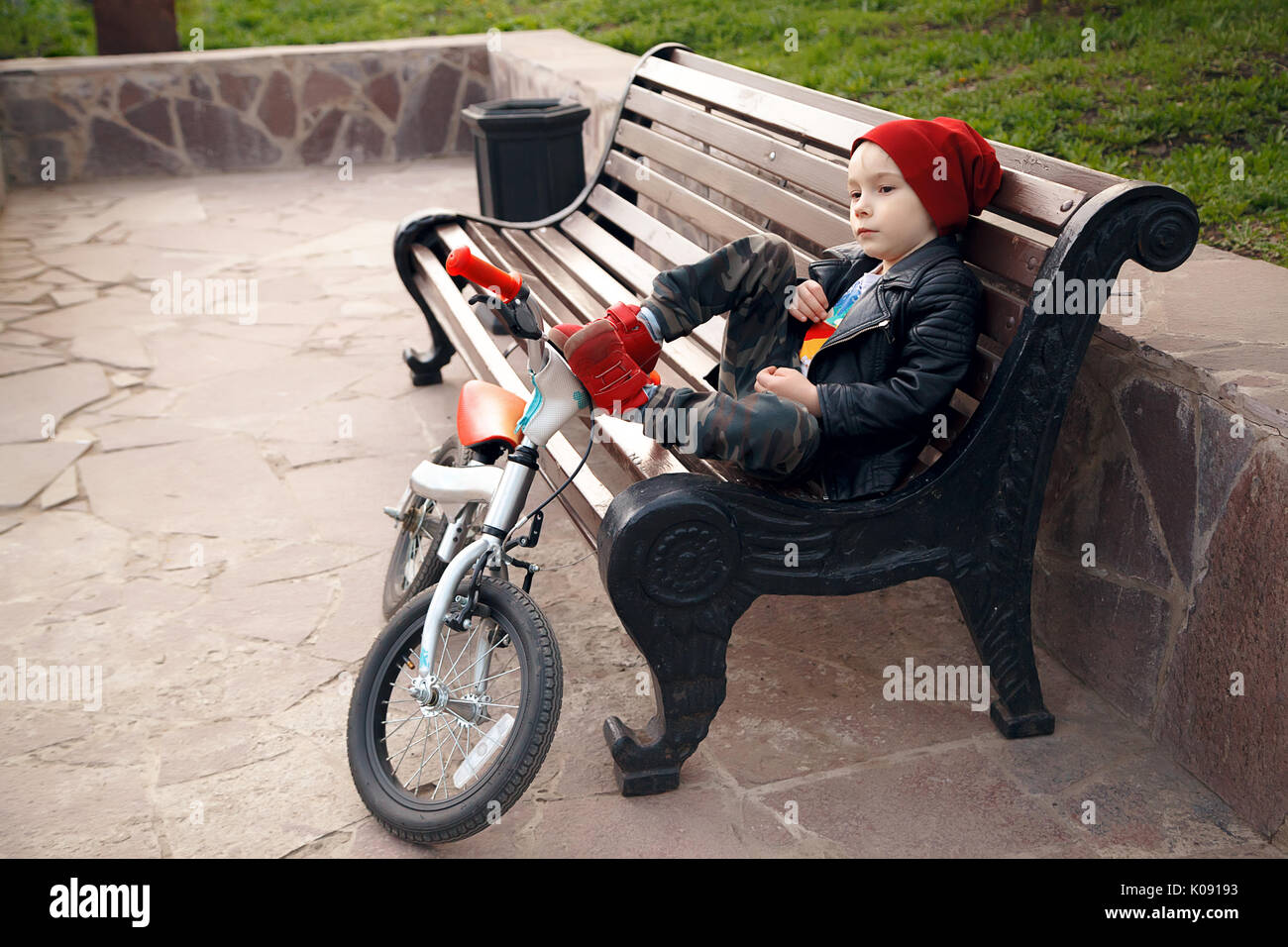 Ritratto di un simpatico ragazzo in un cappuccio rosso-cap, seduta su una panchina in un parco della città con la sua bicicletta e guardando in lontananza. Riposo dopo una bicicletta. Bambino all'esterno. Emozione la fatica Foto Stock