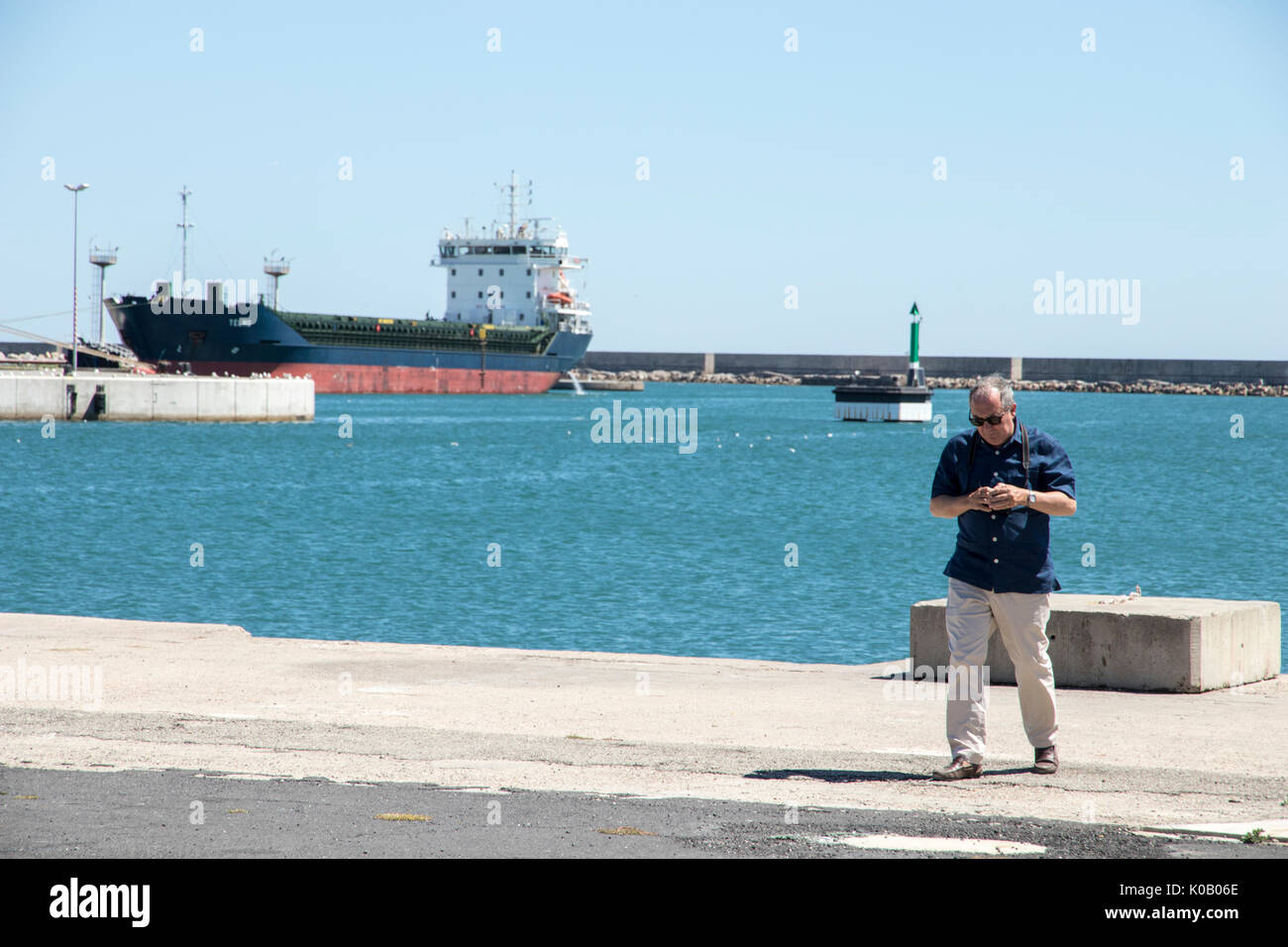 Un fotografo senior nel porto di Sete, Francia, con il mare, navi e una petroliera in background Foto Stock