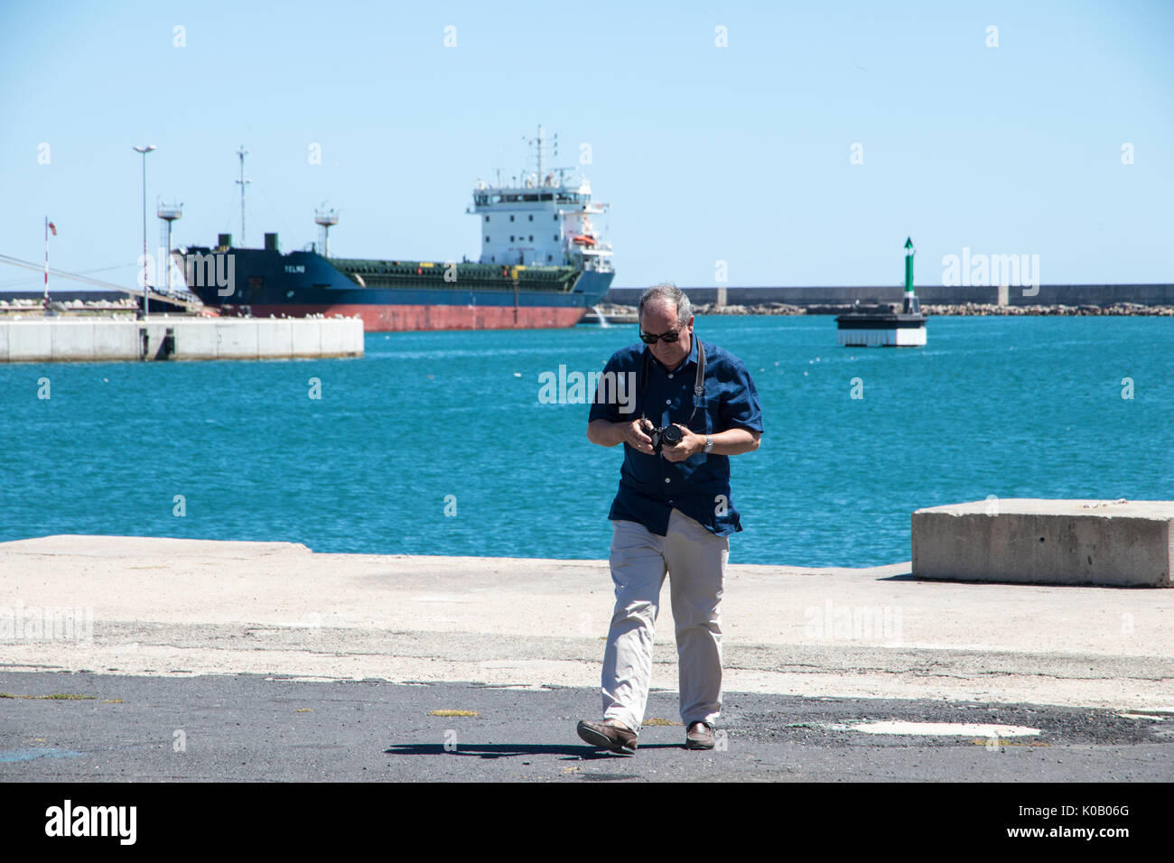 Un fotografo senior nel porto di Sete, Francia, con il mare, navi e una petroliera in background Foto Stock