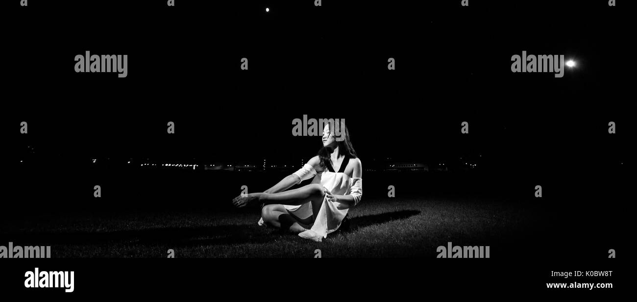 Ballerina in abito bianco si siede sul prato di sera su sfondo di notte le luci. Immagine in bianco e nero. Foto Stock