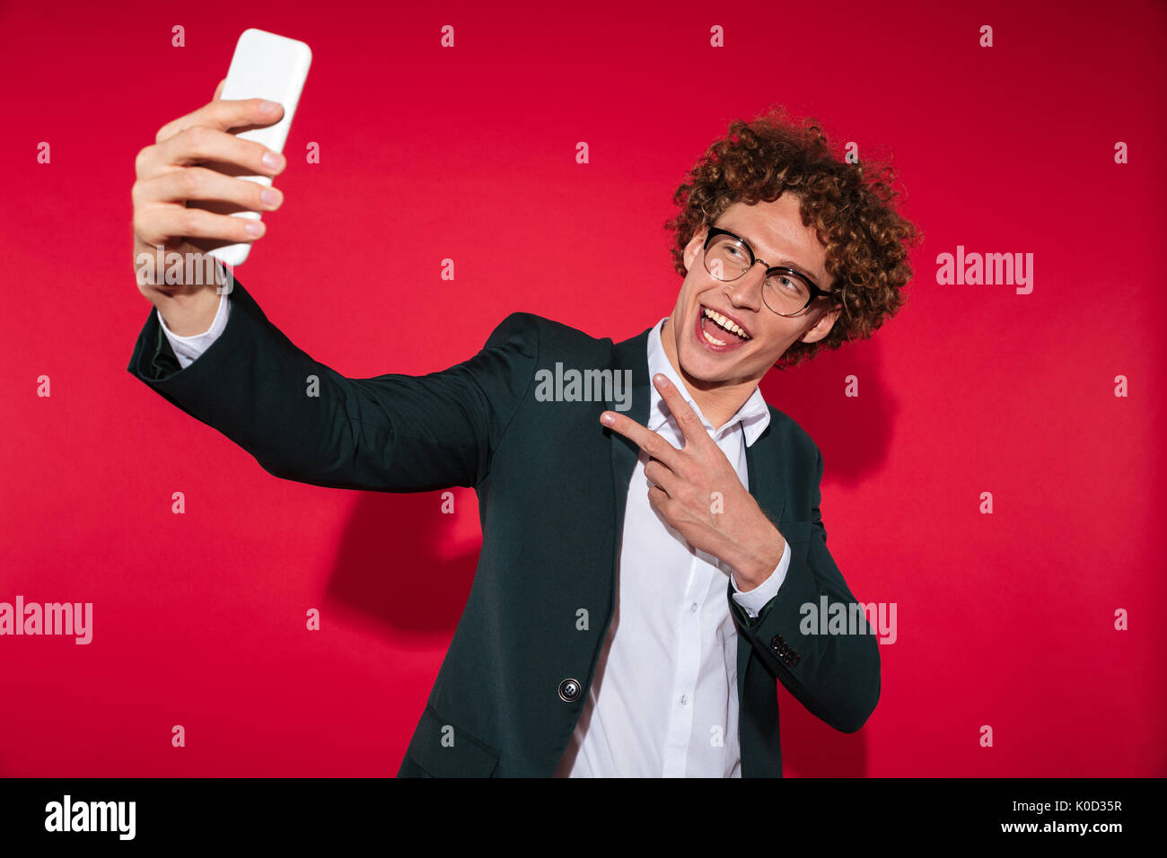 Felice attraente giovane eyewear tenendo selfie e mostra il gesto di pace isolate su sfondo rosso Foto Stock