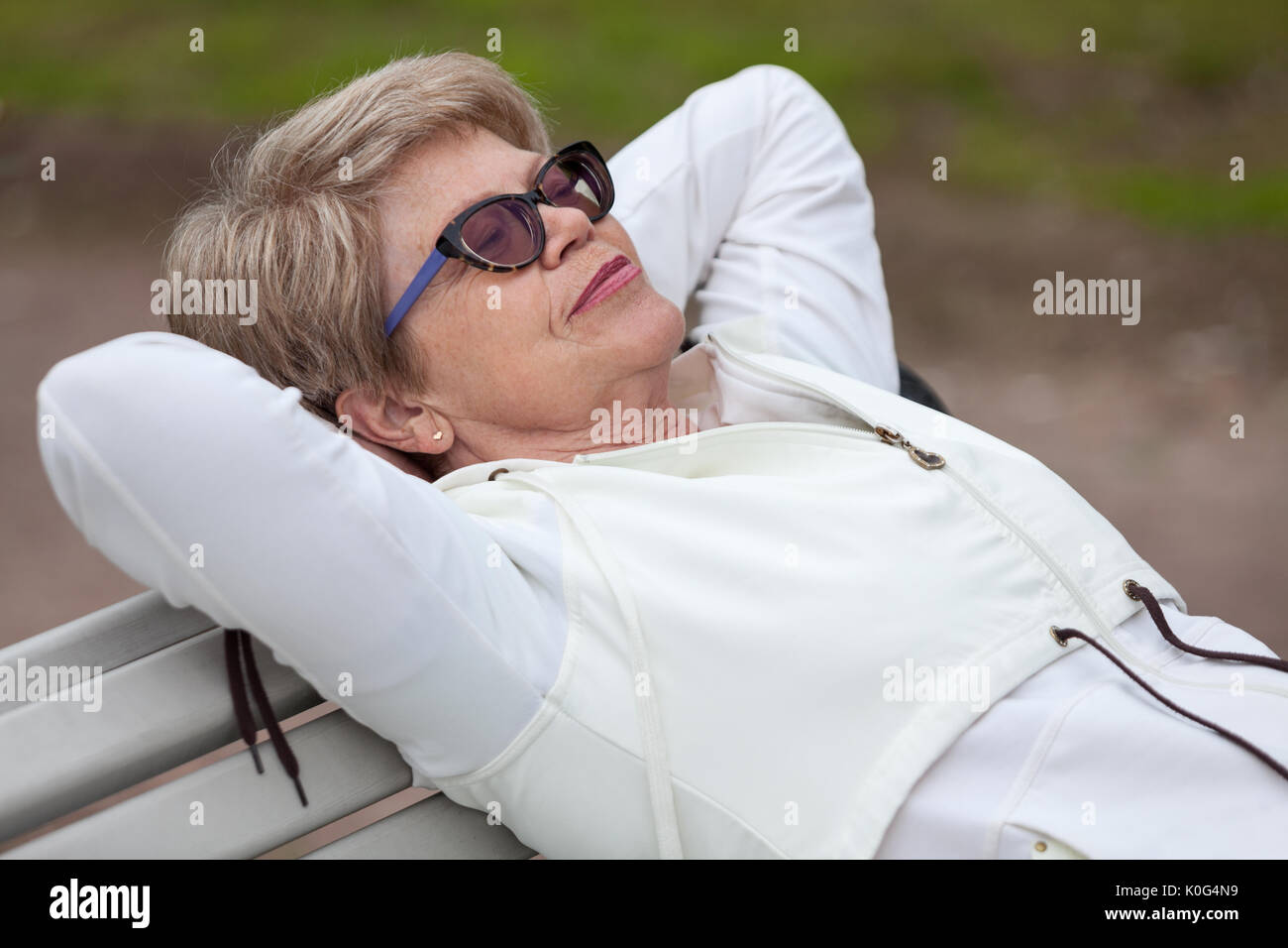 Un anziano donna in bicchieri di dormire sulla sua schiena sul banco in estate park Foto Stock