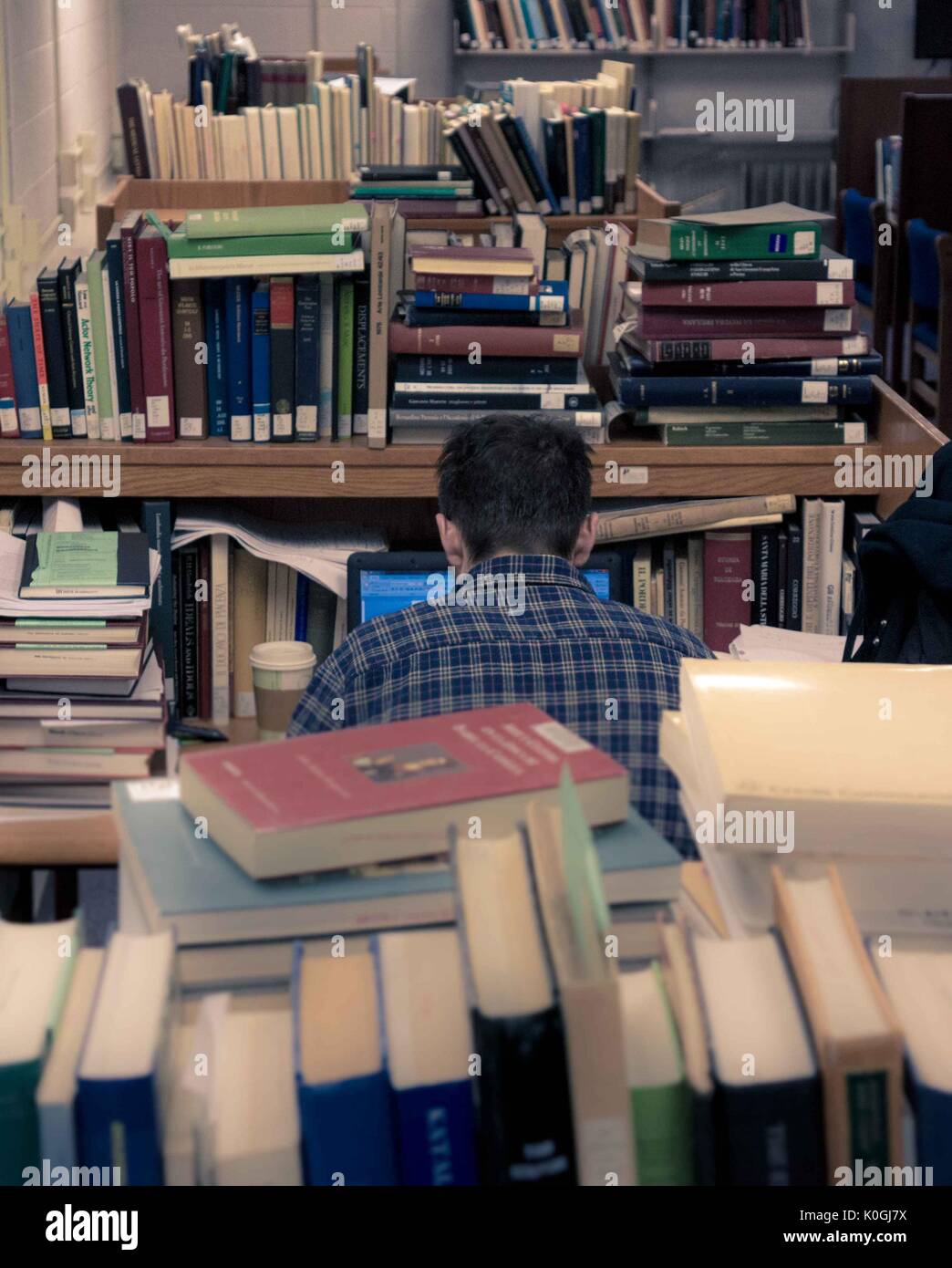 Uno studente laureato si trova nella sua stazione di lavoro sul suo laptop, circondato da mucchi di libri, nella Milton S. Eisenhower Library nel campus Homewood della Johns Hopkins University di Baltimora, Maryland, 2015. Cortesia Eric Chen. Foto Stock