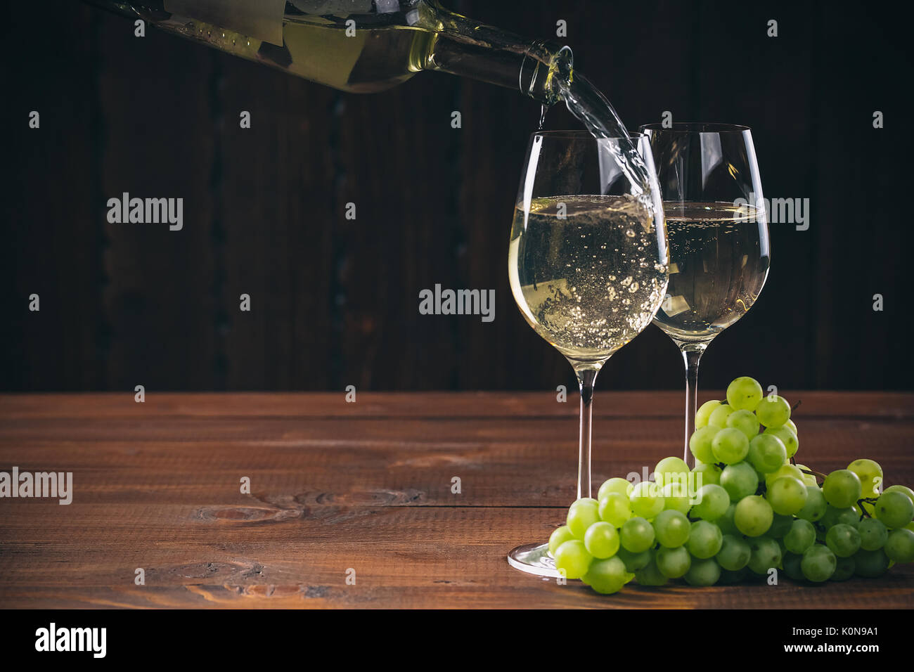 Versare il vino bianco dalla bottiglia in un bicchiere con un grappolo di uva verde contro lo sfondo di legno con spazio libero Foto Stock