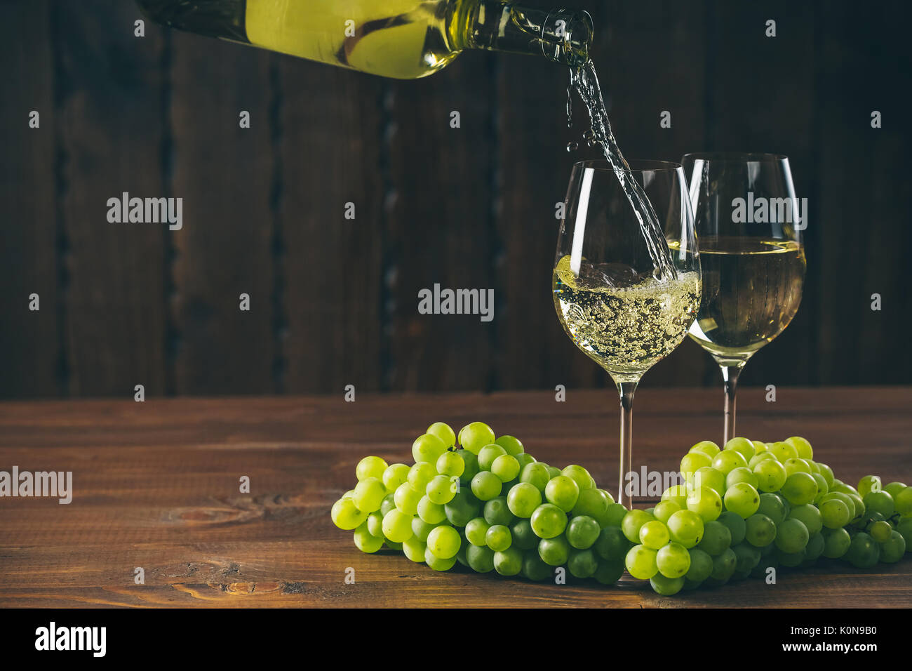 Versare il vino bianco dalla bottiglia in un bicchiere con un grappolo di uva verde contro lo sfondo di legno con spazio libero Foto Stock