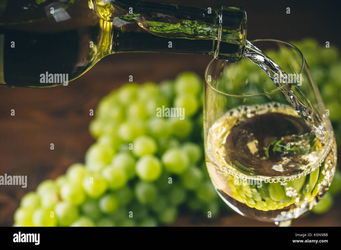 Versare il vino bianco in un bicchiere con un grappolo di uva verde contro lo sfondo di legno Foto Stock