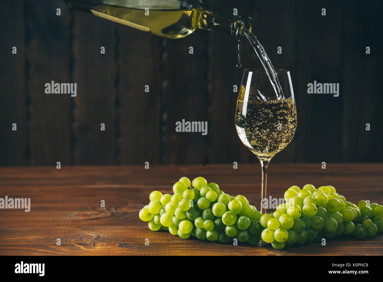 Versare il vino bianco dalla bottiglia in un bicchiere con un grappolo di uva verde contro lo sfondo di legno con spazio libero Foto Stock