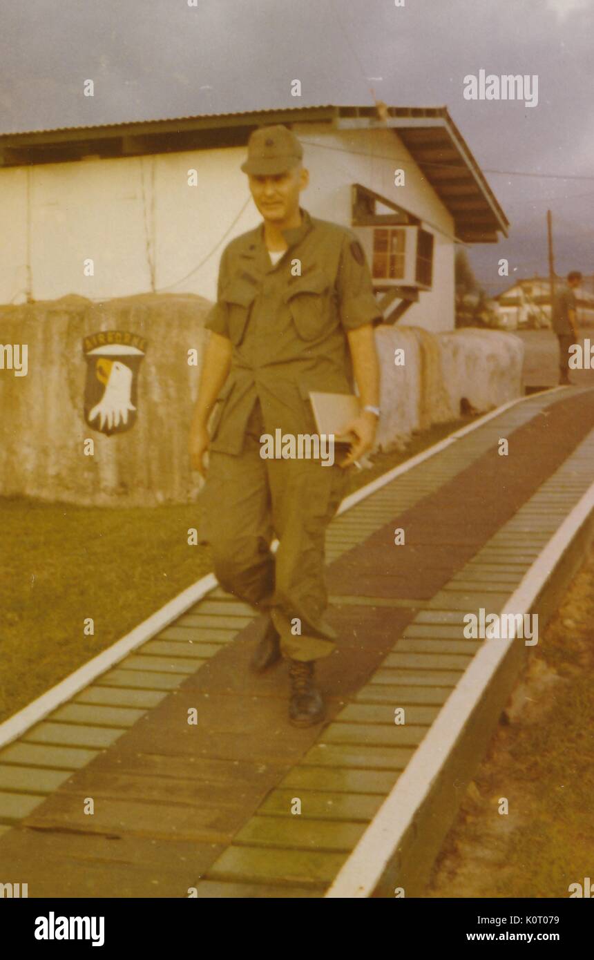Un soldato, identificato come Robert Swisher del 101st Divisione Airborn, può essere veduto camminare verso il basso una passerella in legno, esterno di un edificio con un condizionatore di aria può essere visto con un altro soldato in background, Camp Eagle, Vietnam, 1964. Foto Stock