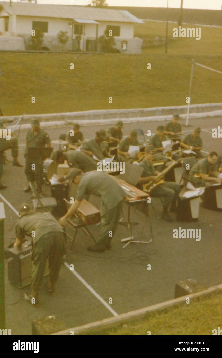 Un gruppo di più di una dozzina di soldati, membri della 101ª Divisione aviotrasportata, imposta per eseguire come una band, consistente di ottoni, percussioni e organo elettrico che stanno utilizzando un campo da pallavolo come il loro spazio per prestazioni, Camp Eagle, Vietnam, 1964. Foto Stock