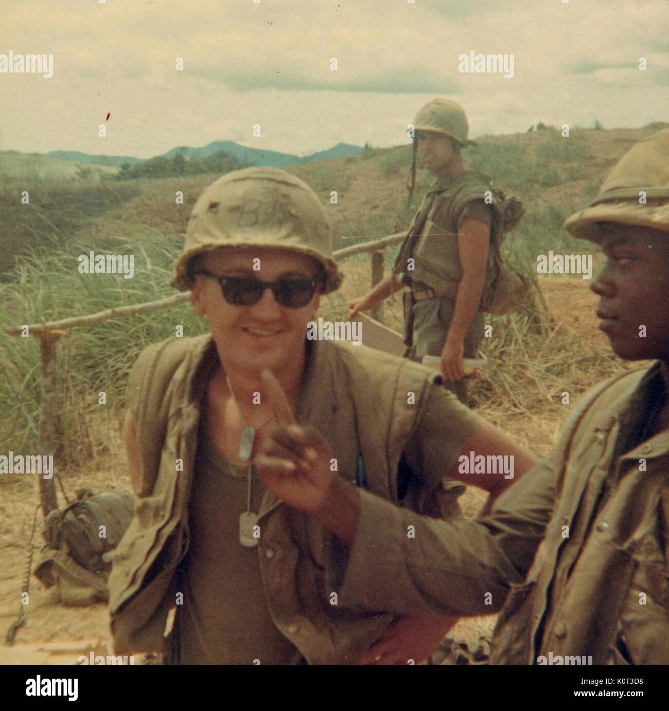 Marine in giubbotto di combattimento e casco, occhiali da sole e dog tag, stando in piedi in un campo durante la guerra del Vietnam, un afro-americano di compagni di Marine rivolta verso di lui come un terzo Marine sorge in background, 1965. Foto Stock