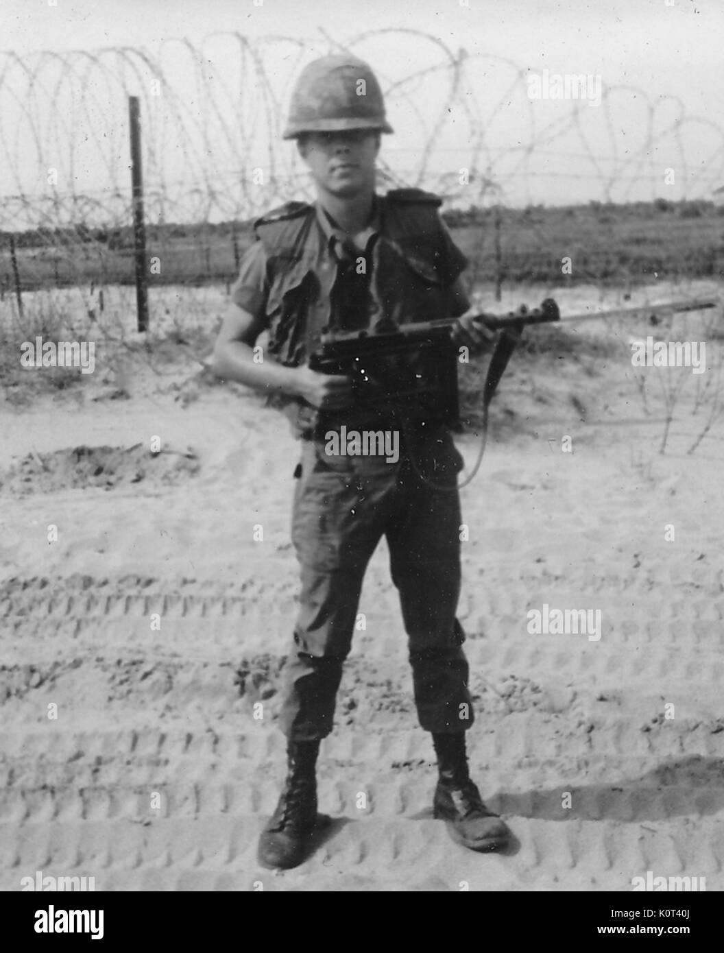 Gli Stati Uniti soldato indossando la sua uniforme di combattimento e casco tenendo un arma, egli è in piedi su le tracce lasciate da un grande veicolo militare, reticolati di filo spinato può essere visto in background, Vietnam, 1967. Foto Stock