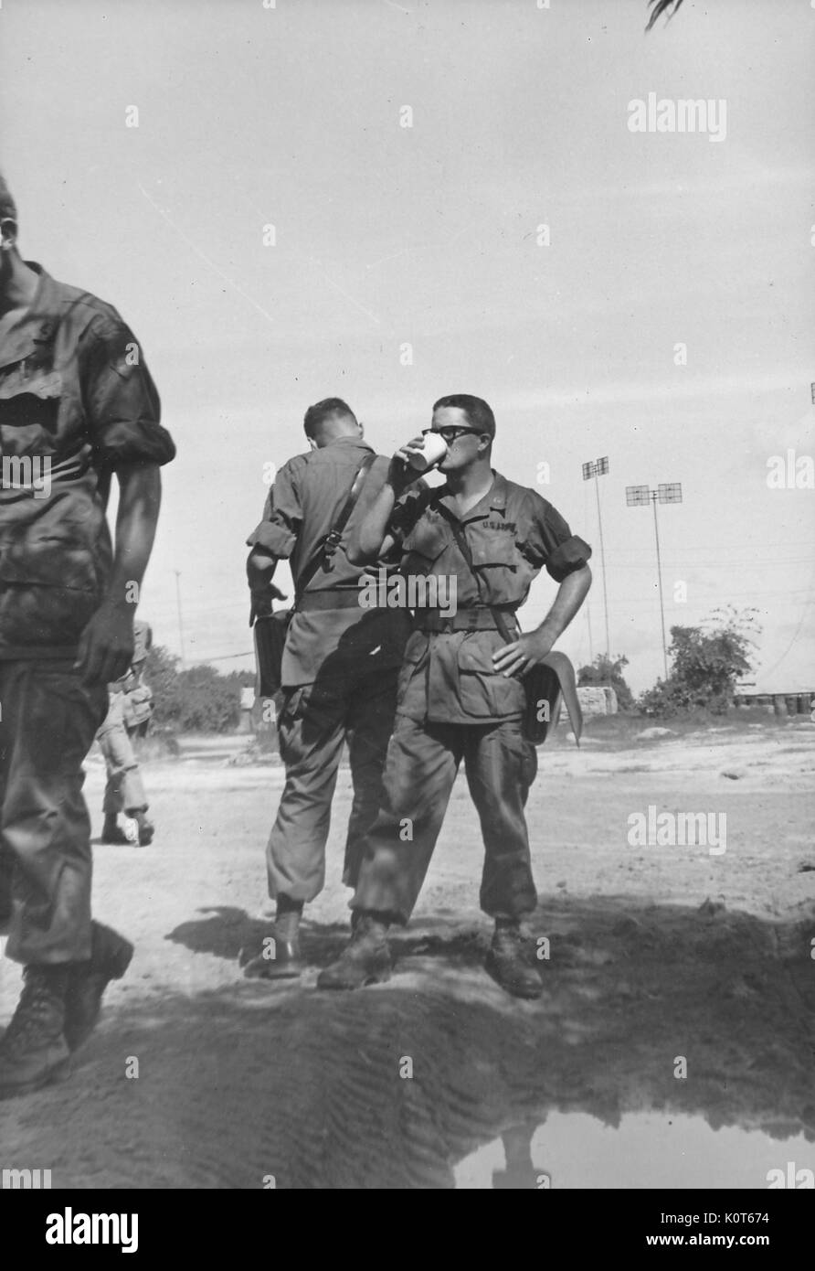 Un piccolo gruppo di soldati riuniti al di fuori, un soldato è di bere da un bicchiere di carta, gli edifici e i grandi antenne direzionali possono essere visti sullo sfondo, Vietnam, 1967. Foto Stock