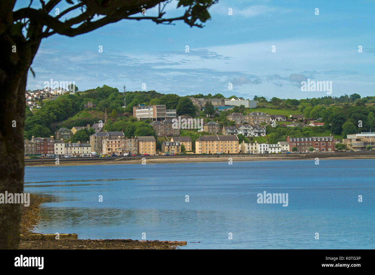 Edifici di fronte mare e collina adiacente alla città di Port Bannatyne accanto calme acque blu dell'oceano sotto il cielo blu sull isola di Bute, Scozia Foto Stock