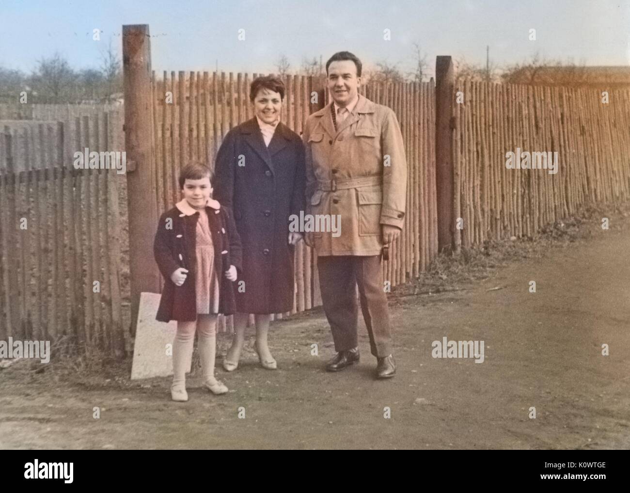 Una famiglia di tre persone su una strada, uomo, donna e bambina vestito di cappotti, in piedi da una staccionata in legno, guardando la telecamera, Germania, 1946. NOTA: l'immagine è stato colorizzato digitalmente usando un processo moderno. i colori potrebbero non essere periodo-precisa. Foto Stock