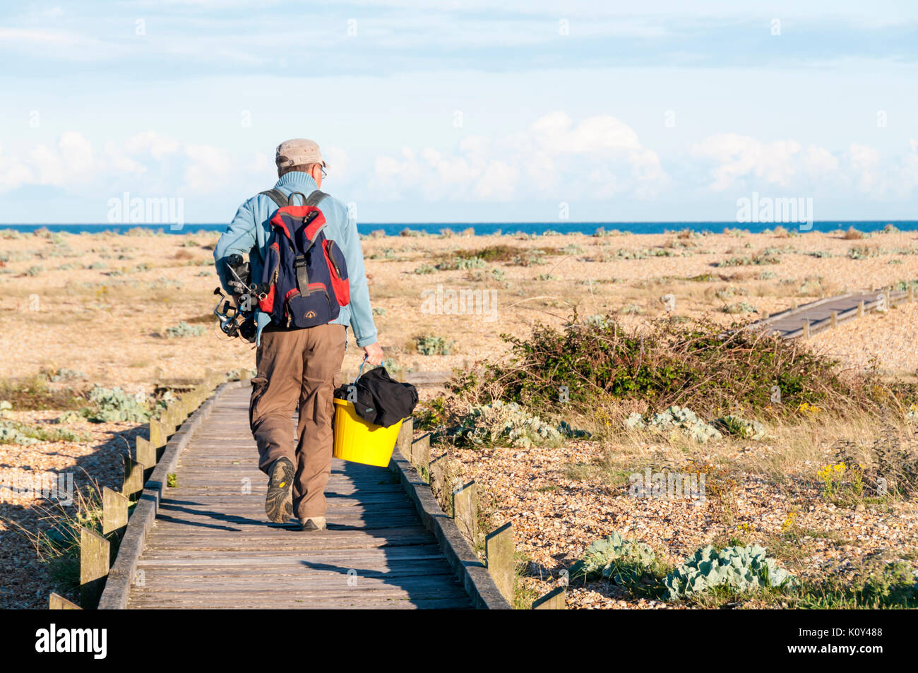 Mare pescatore o il pescatore a piedi lungo una passerella attraverso shingle alla spiaggia di Dungeness nel Kent. Foto Stock