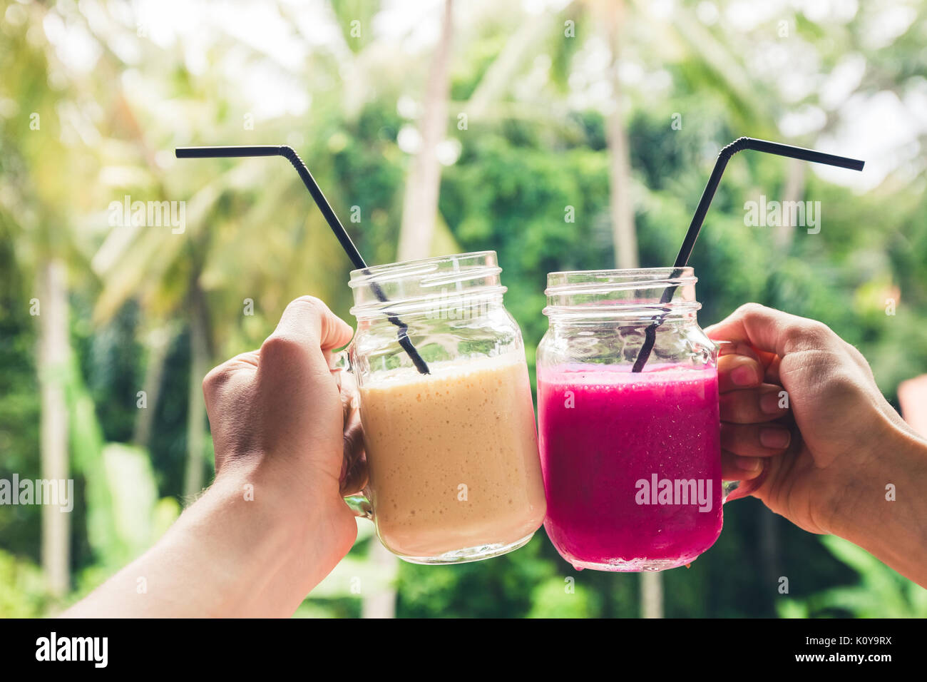 Due coloratissimi frullati di frutta nelle mani. L'estate tropicale e l'umore. Il freddo del bere miscelato, banana e dragon frullato di frutta. Clink occhiali da giovane le mani Foto Stock