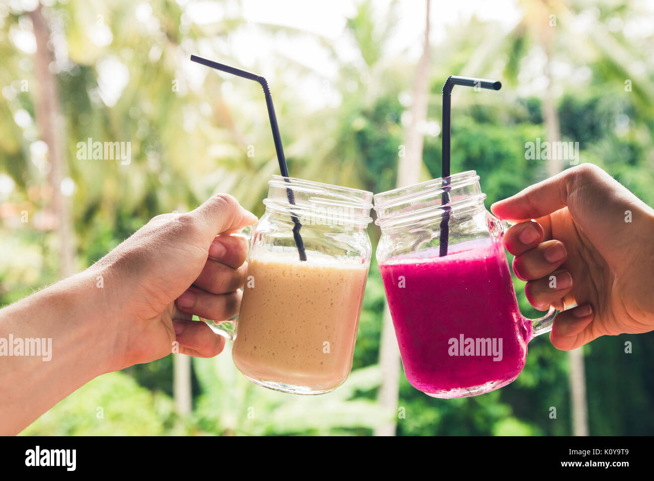 Due coloratissimi frullati di frutta nelle mani. L'estate tropicale e l'umore. Il freddo del bere miscelato, banana e dragon frullato di frutta. Clink occhiali da giovane le mani Foto Stock