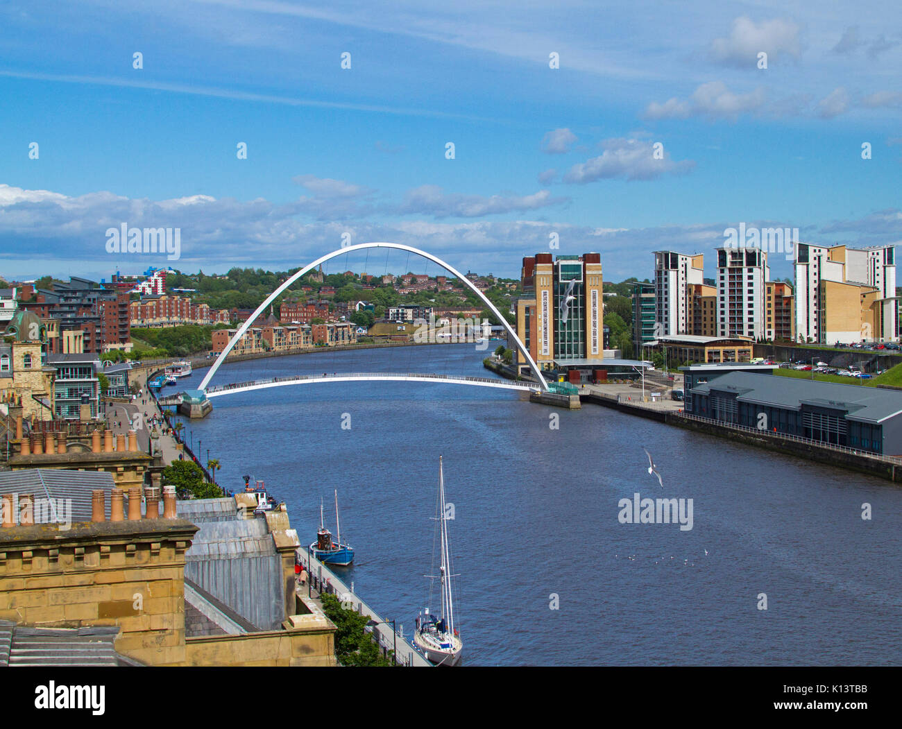 Gateshead Millennium Bridge, di ribaltamento del ponte pedonale di fronte fiume Tyne a Newcastle-upon-Tyne con edifici della città nelle vicinanze sotto il cielo blu Foto Stock