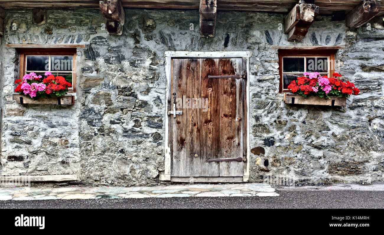 Vecchia fattoria casa fatta di pietre di campo con porta di legno, due finestre e rosso brillante fiori Foto Stock