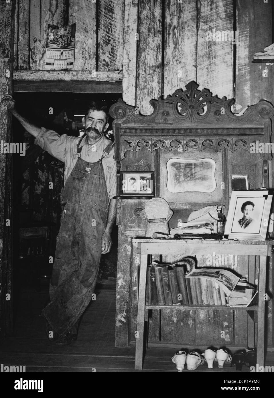 Un sincero a piena lunghezza Ritratto di un agricoltore appoggiata in una porta all'interno di casa sua, dotate di fotografie di famiglia, una piccola collezione di libri e diverse coppie di sua moglie scarpe, 1938. Dalla Biblioteca Pubblica di New York. Foto Stock