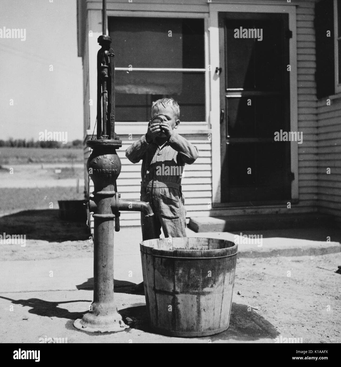 Ragazzo che indossa un manicotto lungo lavoro ponticello, in piedi accanto a una pompa per acqua e di bere da un bicchiere, di fronte a una casa, Fairbury, Nebraska, 1936. Dalla Biblioteca Pubblica di New York. Foto Stock
