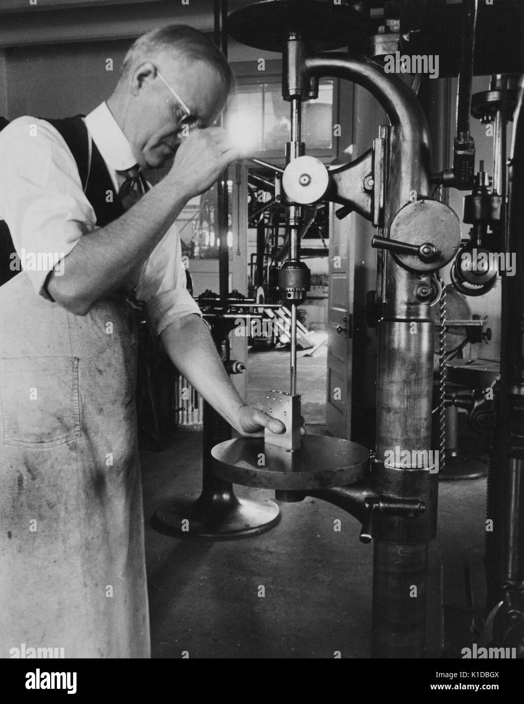 Uomo che indossa un grembiule usurato e utilizza una pressa per trapano per perforare un blocco di metallo in un negozio di macchine, Greenbelt, Maryland, 1935. Dalla Biblioteca pubblica di New York. Foto Stock