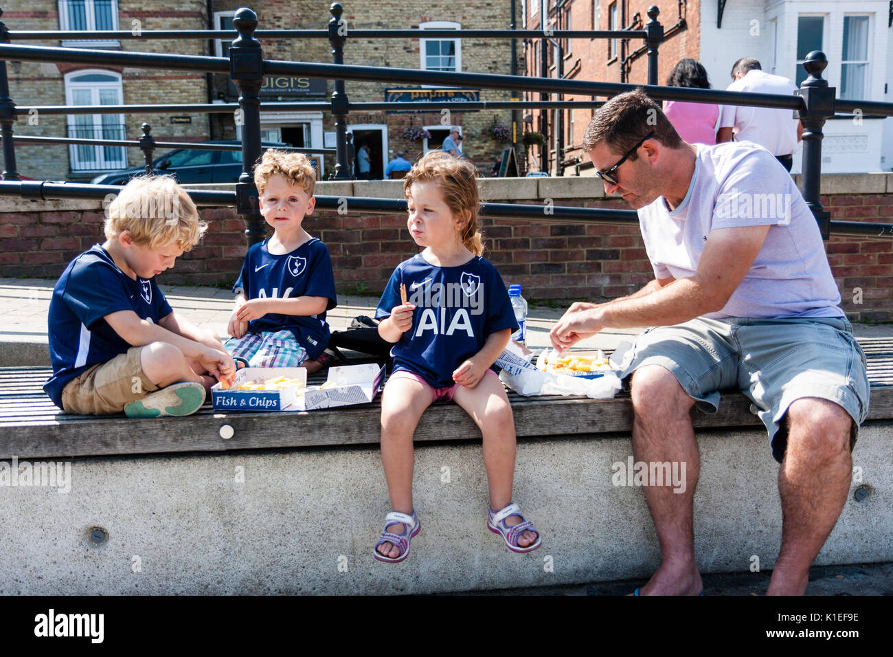 Inghilterra, sandwich. Tre giovani bambini 5-6 anni, con il loro padre, seduto sulla parete condividendo una scatola di pesce e patatine in estate sole brillante. Foto Stock