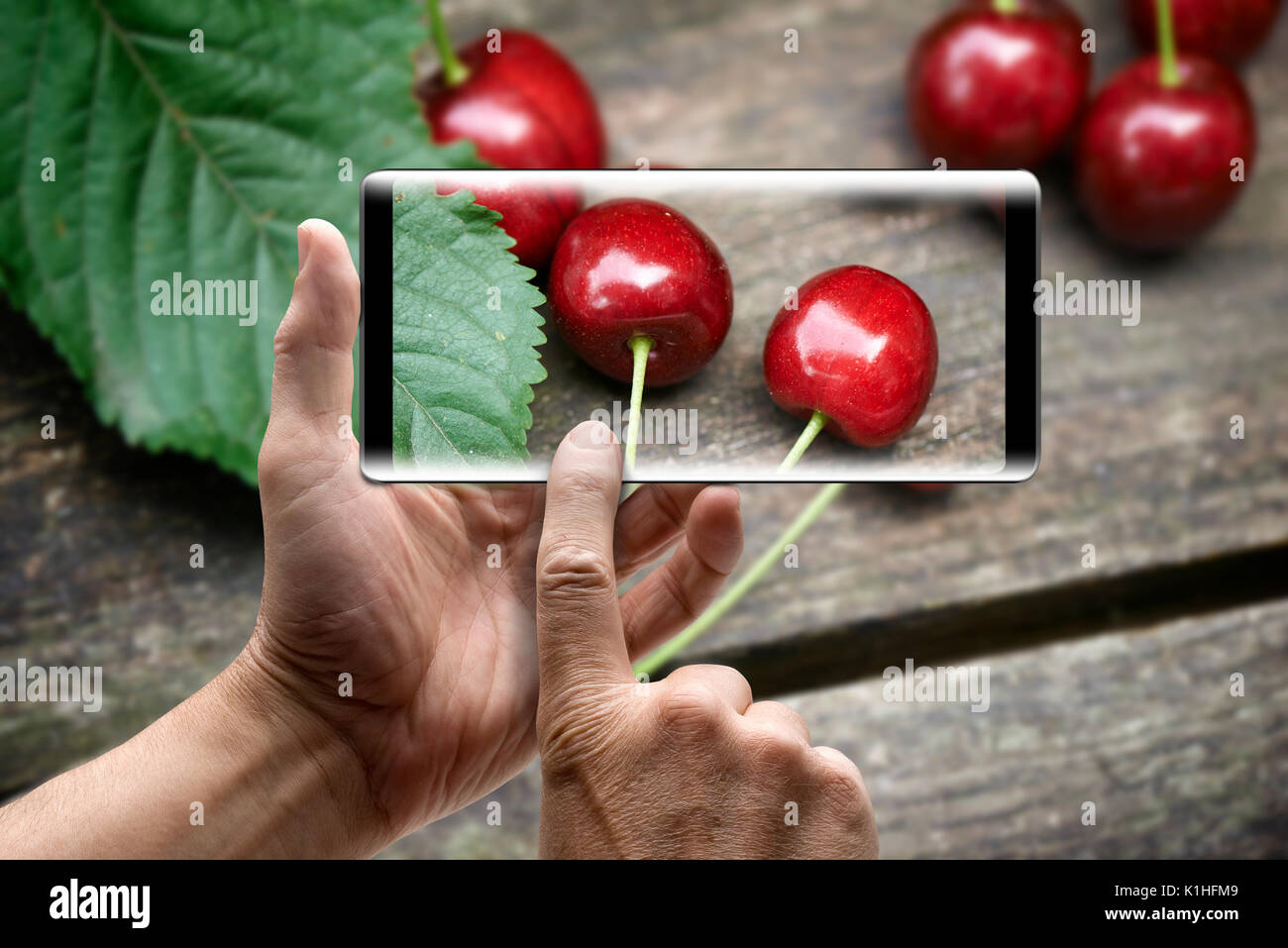 Due mani tenendo un mobile Smartphone e scattare una foto delle ciliege su un tavolo di legno Foto Stock