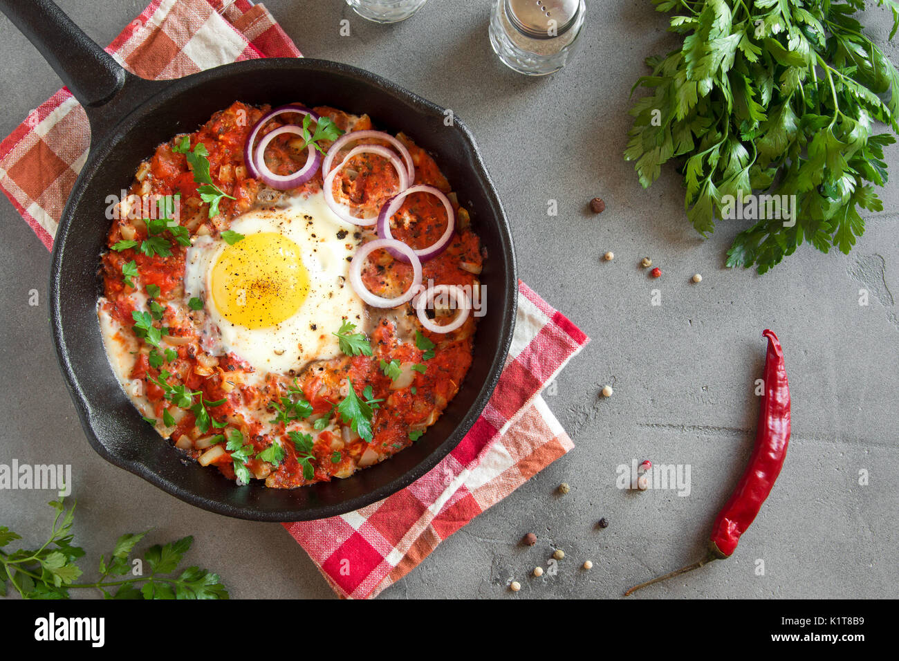 Shakshuka in una ghisa Pan. Stile medio orientale le uova in camicia in salsa di pomodoro e peperone con prezzemolo e cipolla e spezie sul cemento grigio Sfondo. Foto Stock