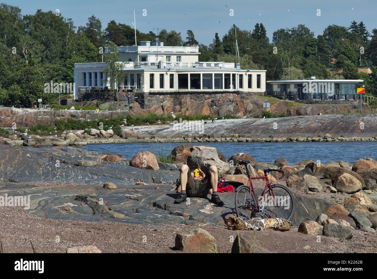 Ciclista rilassante sulla spiaggia rocciosa con grandi massi. Casa con finestre panoramiche in background Foto Stock