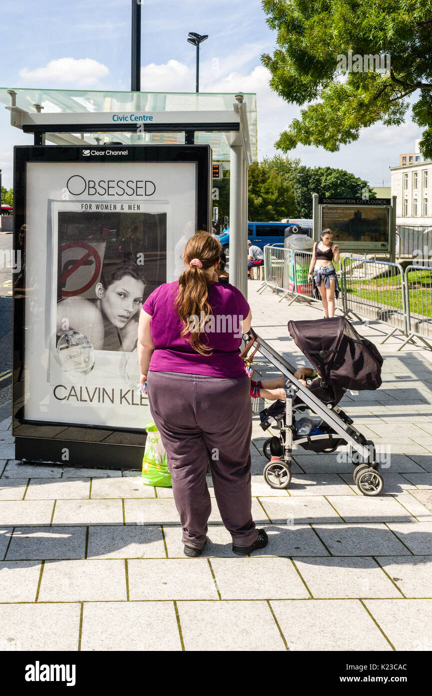 Una grande donna si erge davanti ad una fermata autobus dotato di un annuncio pubblicitario per una marca di profumo illustrato con un modello attraente. È la donna invidioso Foto Stock