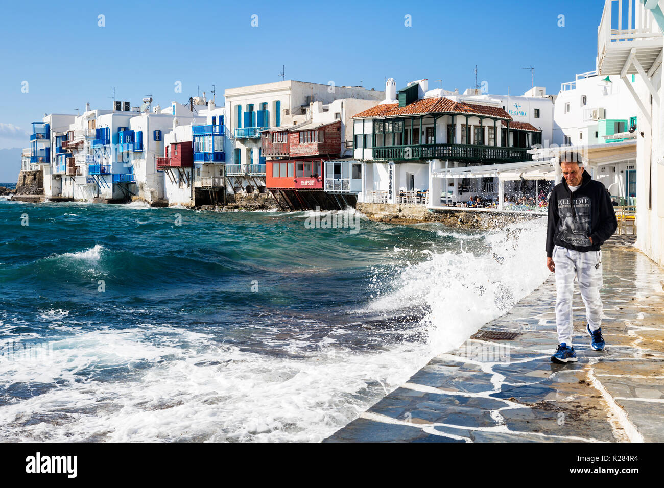 Un uomo che cammina lungo come onde infrangersi contro il marciapiede, Little Venice, Mykonos, Grecia. Foto Stock
