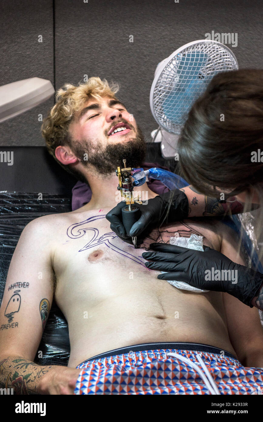 Cornovaglia Tattoo Convention - un cliente smorfie come tattooist tatuaggi un progetto sul suo petto in Cornovaglia Tattoo Convention. Foto Stock