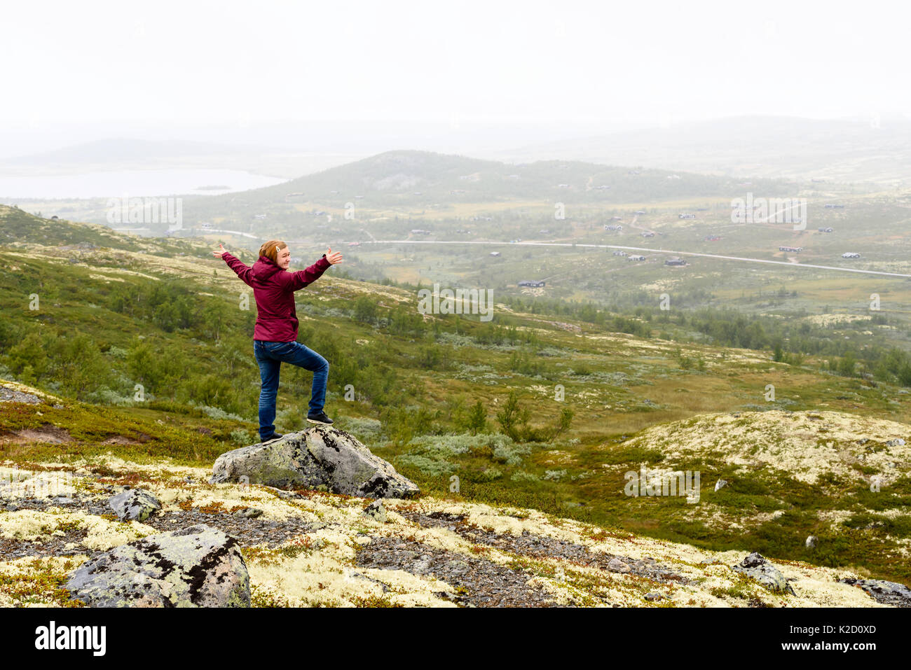 I giovani adulti che si affacciava su foggy mountain view con il piccolo villaggio e strada. Arma fuori e una faccia felice. Ubicazione Vasstulan, Norvegia. Foto Stock