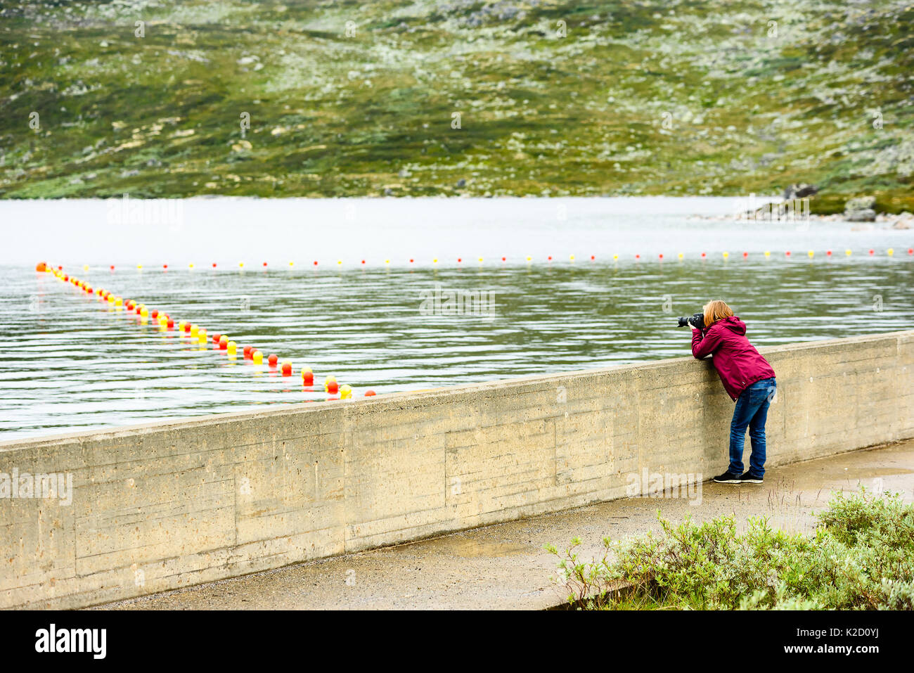 Giovane donna adulta prendendo foto di boe di sicurezza lungo una fune in acqua da una piccola diga. Paesaggio di montagna in background. Foto Stock