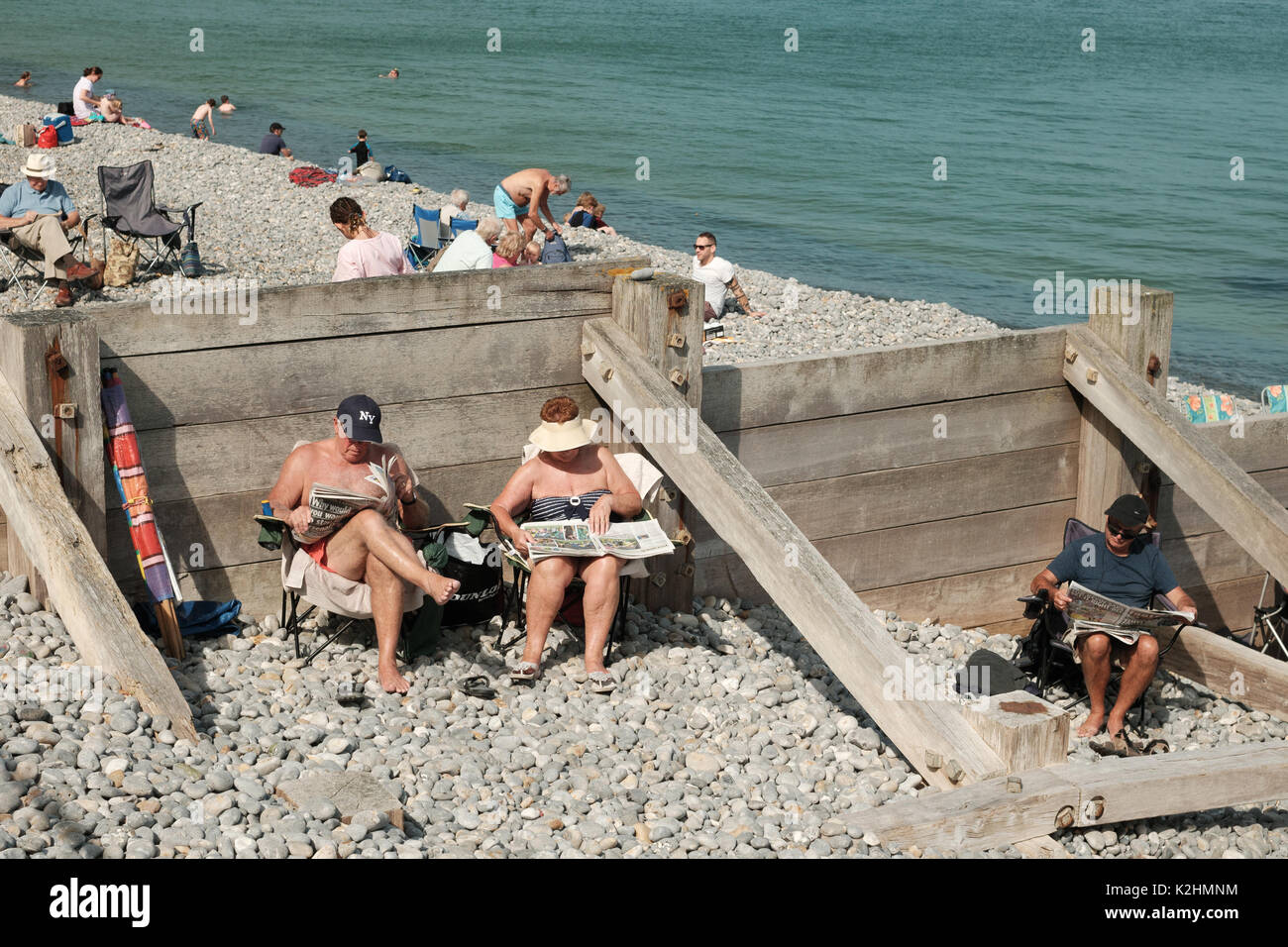 Coppia di mezza età leggere quotidiani in sedie a sdraio sulla spiaggia con ciottoli in Norfolk, Regno Unito, su un caldo weekend festivo Foto Stock