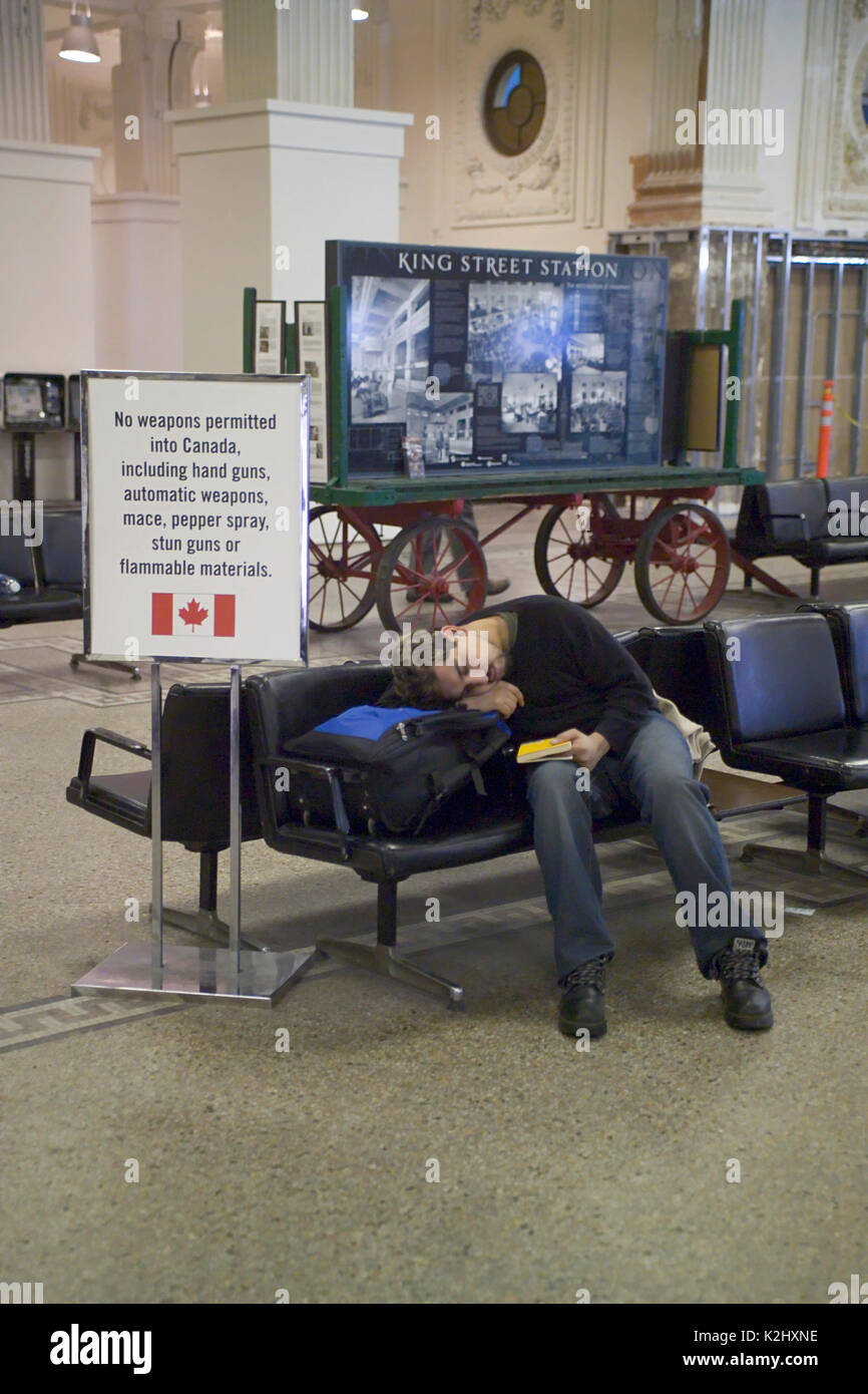 Stanco di un maschio adulto giovane viaggiatore pan su un banco in King Street Stazione Ferroviaria a Seattle, WA, accanto a un cartello di avvertimento di divieti di armi nelle vicinanze Canada. Foto Stock