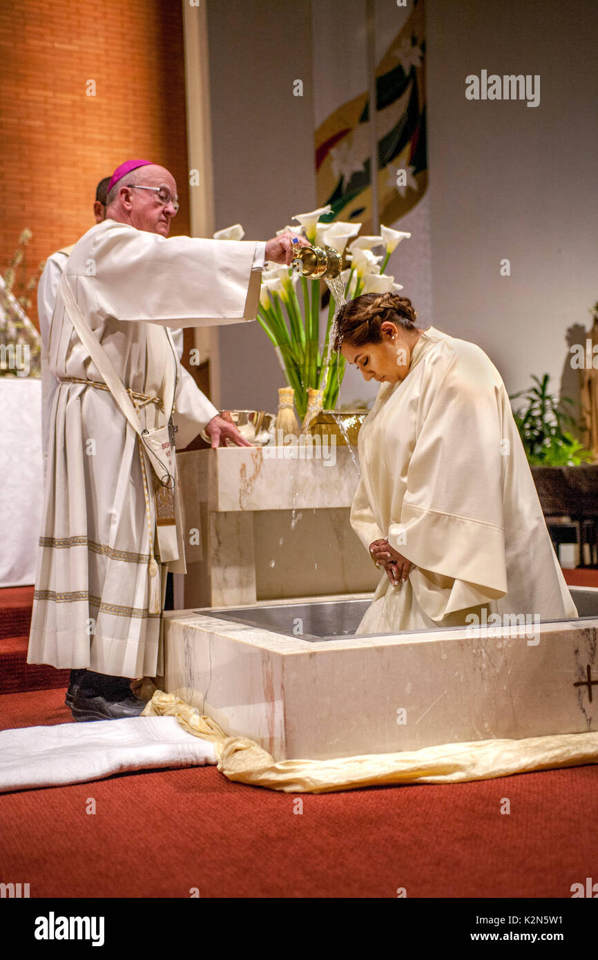 Una donna ispanica che indossa un abito battesimo nel font di una cattedrale cattolica in arancione, CA, centine come un vescovo unge lei con l'acqua santa. Foto Stock