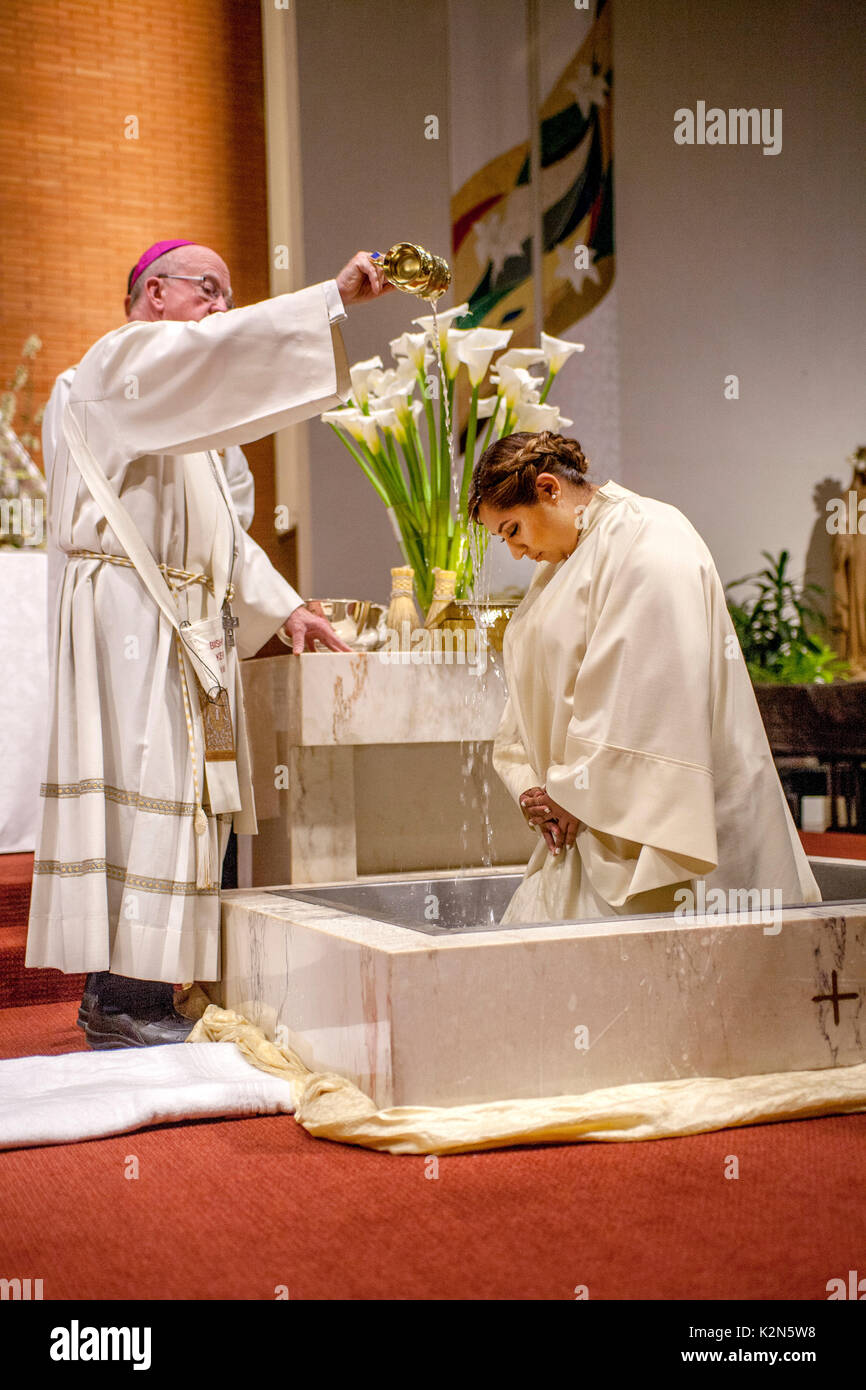 Una donna ispanica che indossa un abito battesimo nel font di una cattedrale cattolica in arancione, CA, centine come un vescovo unge lei con l'acqua santa. Foto Stock