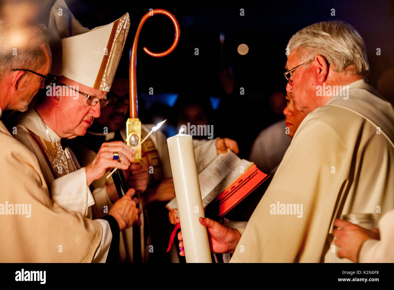 Dalla luce del fuoco pasquale, un vescovo cattolico utilizza un cono di luce di una candela processionale detenute da un diacono derubato prima di condurre una massa di battesimo presso un Orange, CA, Cattedrale. Foto Stock