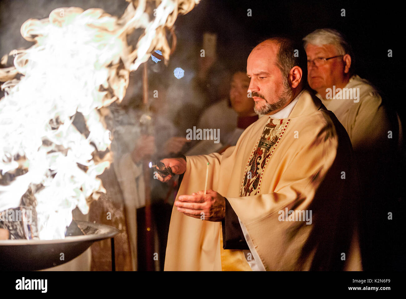 Dalla luce del fuoco pasquale, un sacerdote ammantato accende una conicità alla luce di una candela processionale prima di una massa di battesimo presso un Orange, CA, cattedrale cattolica. Foto Stock