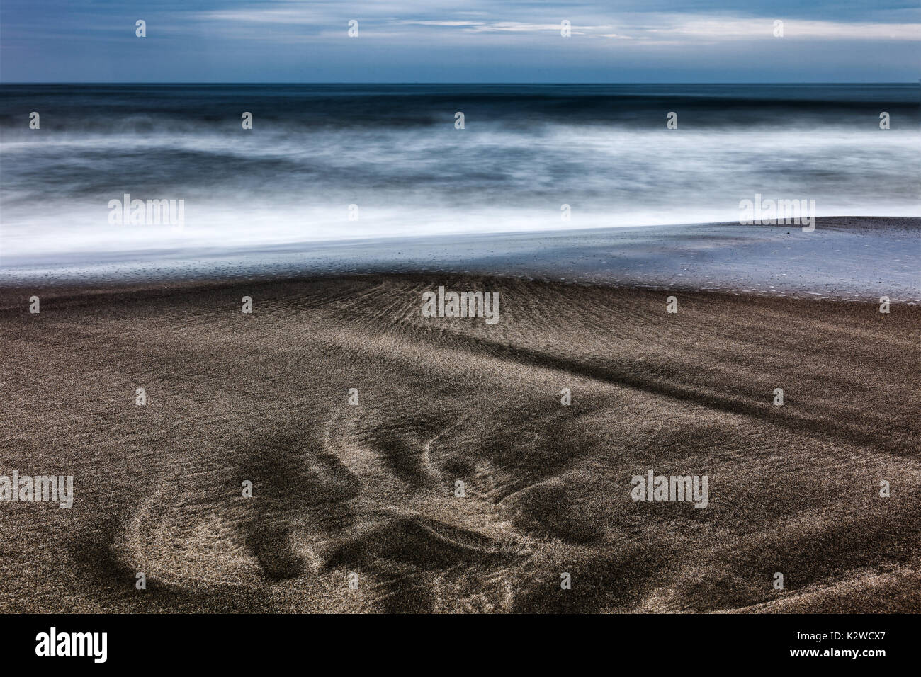 Una misteriosa impronta di sabbia scura evoca le tracce di una creatura preistorica, che si trova contro onde nebbiose e un paesaggio marino lunare e senza tempo. Foto Stock